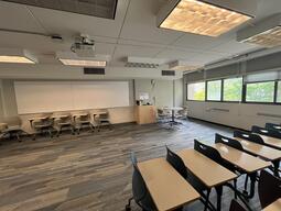 View of the classroom from a student seat near the rear of the room. In front are multiple empty tables and chairs. The instructor’s podium is at the front of the room on the right. A projector on the ceiling projects onto a screen that is mounted on the front wall. This classroom is accessible for students.