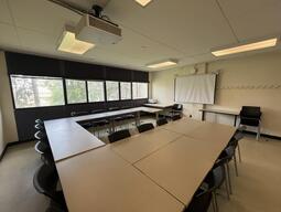 View of the classroom from the front of the classroom. There are multiple tables and chairs for students, arranged in a rectangle in the middle of the room. This classroom is accessible for instructors.