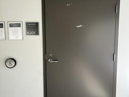 The entrance to room B201 in Mackintosh-Corry Hall is a dark grey door with a lever on the left side and an automatic operator. There is another accessible door for instructor's to access the podium, around the corner.