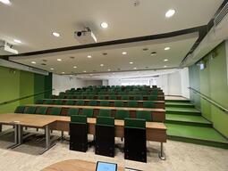 View of the classroom from the podium. The rows of seating have tiers towards the back of the room. There are stairs on either side of the room to access the back tiers. The seats are attached to the desks and there is no power at the tables. At the front of the room are two wheelchairs accessible seats with chairs pulled up to the desks.