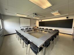 View of the classroom from a student seat near the rear of the room. In front are multiple empty tables and chairs. A long blackboard is located on the front wall. A projector on the ceiling projects onto a screen that extends from the ceiling. The entryway door is located at the front of the room on the right. This classroom is accessible for students.