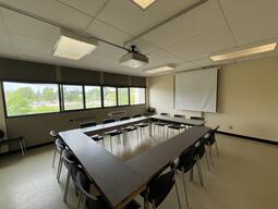 View of the classroom from the front of the room. There are tables and chairs for students, arranged in a rectangle in the center of the room. This classroom is accessible for instructors.