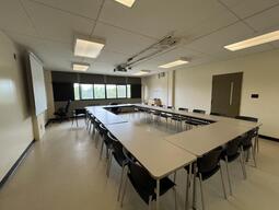 View of the classroom from a student seat near the rear of the room. In front are multiple empty tables and chairs. A long blackboard is located on the front wall. A projector on the ceiling projects onto a screen that extends from the ceiling. This classroom is accessible for students.