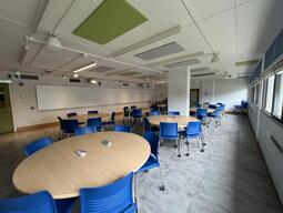View of the classroom from the student's perspective. In front are several round tables with 4 chairs each. Each table has 4 outlets. The projector's on the wall project onto the whiteboards around the room. This classroom is accessible for students.