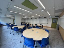 View of the classroom from the student's perspective. In front are several round tables with 4 chairs each. Each table has 4 outlets. The projector's on the wall project onto the whiteboards around the room. This classroom is accessible for students.