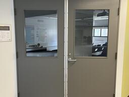 The entryway door of room D214 in Mackintosh Corry Hall is a double grey door with a lever handle on the right door.