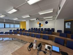 View of the classroom from the instructor’s podium. There are rows of long fixed tables and blue chairs that swivel from the table. The wall at the back of the room has areas of yellow paint and colorful hexagon panels. This classroom has tiered steps to access the middle and back areas of the room. The entryway door is located at the back of the room.