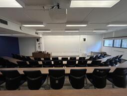 View of the classroom from a student seat near the rear of the room. In front are rows of wood tables and blue chairs. The instructor’s podium is at the front of the room on the left. The front wall of the classroom is covered in a whiteboard surface. A digital projector on the ceiling projects onto the whiteboard surface at the front of the classroom.