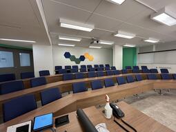 Instructor’s view of the desks throughout the room. There are four rows of seats on tiers raising towards the back of the room. There is wall art on the back wall of the room. Chairs and tables are stationary with the chairs attached to the desks.