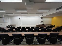 Student’s view while sitting in the classroom. There are two rows of seats facing the front of the class as well as the podium at the front right of the room. When presenting media images are projected on the front wall of the room which is also a whiteboard wall.