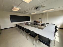 View of the classroom from a student seat near the rear of the room. In front are multiple empty tables and chairs. A long blackboard is located on the front wall. The entryway door is located at the front of the room on the left. This classroom is accessible for students.