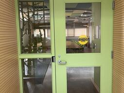 Door to enter room E202 in Mackintosh-Corry Hall. The door is green with a lever on the left side. There is an automatic operator.