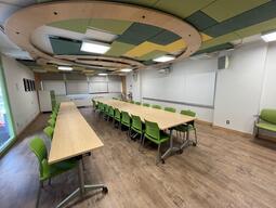 View of the classroom from near the rear of the room. In front are multiple empty tables and chairs. A long whiteboard is located on the front wall. A projector on the ceiling projects onto a screen that extends from the ceiling. The entryway door is located on the right side of the classroom. This classroom is accessible for students.