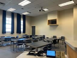 View from the back of the room: Narrow moveable tables with standard  moveable chairs set up in rows facing the podium.