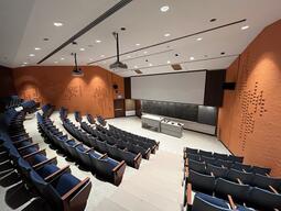 View of the classroom from a student seat near the rear of the room. In front are multiple rows of empty chairs. The instructor podium is at the front of the room, in the middle. The classroom has a long blackboard in the middle of the front wall. A digital projector on the ceiling, projects onto a screen at the front of the room. The entryway door is located at the front of the room, on the left. This classroom is not accessible for students.