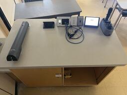 Top-down view of the instructor’s podium. On the podium is a document camera and the Kramer ring equipped with a variety of connections compatible with different computers. There are controls for the lights as well as two microphones on a charging stand.