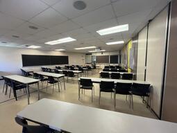 View of the classroom from a student seat near the rear of the room. In front are multiple rows of empty tables and chairs. The instructor podium is at the front of the room on the left. The classroom has a blackboard in the middle of the front wall. A digital projector on the ceiling, projects onto a screen that extends from the ceiling. The entryway door is located at the front of the room, on the left. This classroom is accessible for students.