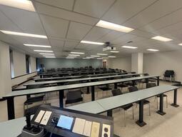 The room from the instructor’s podium. Tables are in rows and are not moveable. Chairs are moveable and are lined in rows behind the desks, facing the front of the room. There is a set of stairs at the back of the room leading to a second exit door. The room is tiered towards the back of the room.