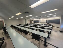 Students view while sitting in the classroom. The first four rows of tables are visible with chairs at the tables, facing the front of the room. The front of the room is a wall of blackboards and the instructors podium is at the front right side of the room.