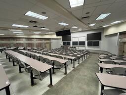 View of the classroom from a student seat near the rear of the room. In front are multiple empty chairs. The instructor podium is at the front left. There are 6 blackboards, and 2 video wall displays at the front of the room.