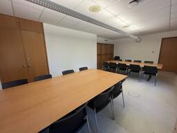 View of the classroom from the front of the room. There are multiple chairs for students, around a large table in the centre of the room. The entryway door is located at the back of the room, on the right. This classroom is accessible for instructors.