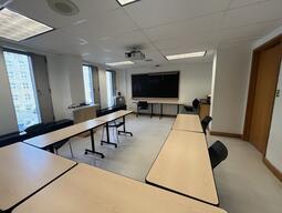 View of the classroom from a student seat near the front of the room. In front are multiple empty chairs and tables. The instructor podium is located on the right side of the classroom. A digital projector on the ceiling, projects onto a screen that extends from the ceiling. This classroom is accessible for students.