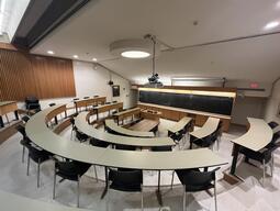 View of the classroom from a student seat near the rear of the room. In front are multiple rows of empty tables and chairs. The instructor’s podium is located at the front of the room on the left. A long blackboard is located on the front wall of the classroom. A digital projector on the ceiling, projects onto a screen that extends from the ceiling. This classroom is not accessible for students.