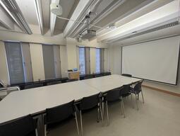 View of the classroom from a student seat near the rear of the room. In front are multiple empty chairs around a large table. The instructor’s podium is located at the front of the room on the right. A blackboard is located on the front wall of the room. This classroom is accessible for students.