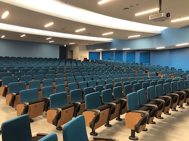 Large tiered auditorium classroom with dark blue chairs and blue painted walls.