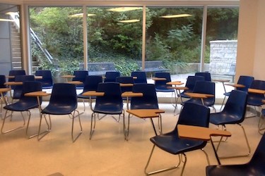 A classroom with many blue chairs with wood tablets for writing. Behind the chairs is a wall of windows looking into a courtyard.