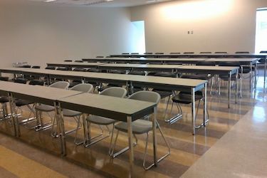 Flat classroom with white walls, rows of grey movable tables and chairs.