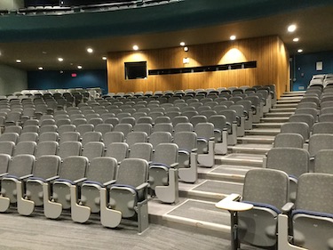 Tiered classroom with grey seats. Each seat has a grey tablet writing surface. Room is dimly lit with spot lights at the back of the class.