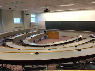 View from the top of a tiered classroom. Desks and chairs arranges in U-shape. A podium and large blackboard along the front wall.