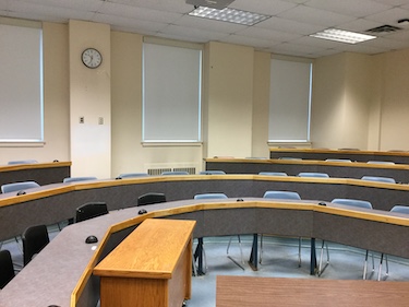 A classroom with desks in an arc shape and loose blue chairs. The room has beige walls and windows with blinds lowered.