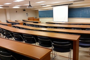 Tiered classroom with brown tables and loose chairs. The front of the room has a long blackboard and a white screen the is lowered in front of the blackboard.