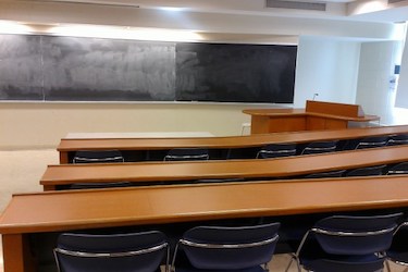 Classroom with rows of brown wood desks and loose chairs. At the front of the room is a long blackboard in podium for the instructor.