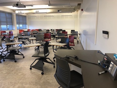 Classroom with many chairs on casters, each with a tablet arm writing surface. Whiteboards on the walls.