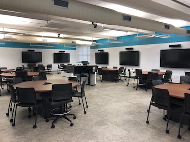 Classroom with round tables with chairs and large televisions mounted on the wall.