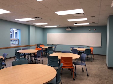 Round tables with moveable chairs. The walls are a pale blue and there is a whiteboard along the back of the room. 