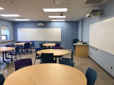 Round tables with moveable chairs. The walls are a pale blue and there is a whiteboard along the back and side of the room.