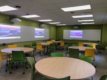 Round tables with moveable chairs. The walls are a soft green and there is a whiteboard along the back and side of the room.
