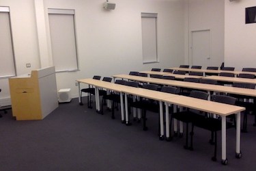 View from the doorway: Narrow moveable tables with standard black moveable chairs set up in rows facing the podium