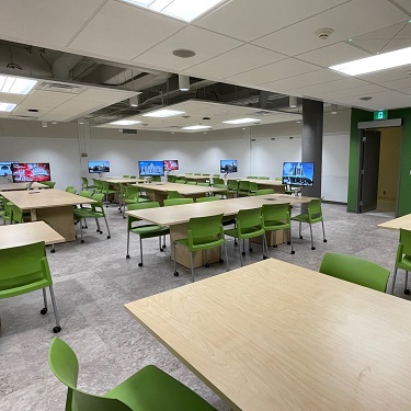 overview of classroom with group tables of 8 with a large monitor at one end and green chairs