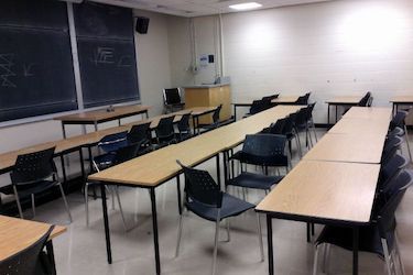 View from the side of the room: Narrow moveable tables with standard black moveable chairs set up in rows facing the podium and blackboard.