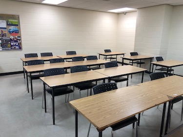 View from the front of the room: Narrow moveable tables with standard black moveable chairs set up in rows facing the front with white cement walls.