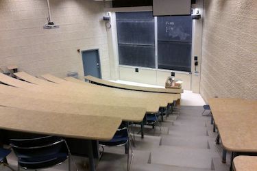View from the back of the room: auditorium with rows of fixed narrow tables with moveable chairs. Rows are at a steep angle.