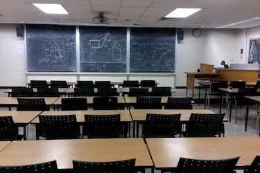 View from the back of the room: Narrow moveable tables with standard black moveable chairs set up in rows facing the podium and blackboard.
