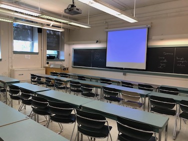 View from the back of the room: Narrow moveable tables with standard  moveable chairs set up in rows facing the podium.