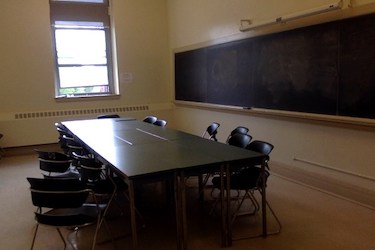 View from the side of the room: Narrow moveable tables with standard  moveable chairs set up in a rectangle with a blackboard on one wall.