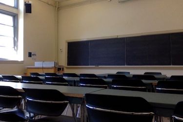 View from the back of the room: Narrow moveable tables with standard  moveable chairs set up in rows facing the podium.